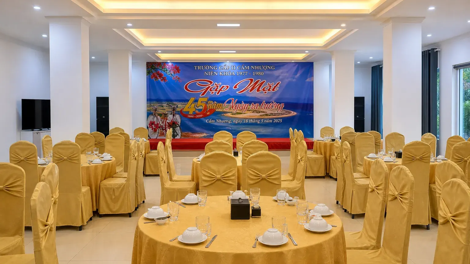 Sảnh tiệc trong nhà với bàn tròn phủ khăn vàng, sân khấu backdrop và hệ thống âm thanh sẵn sàng cho chương trình gala