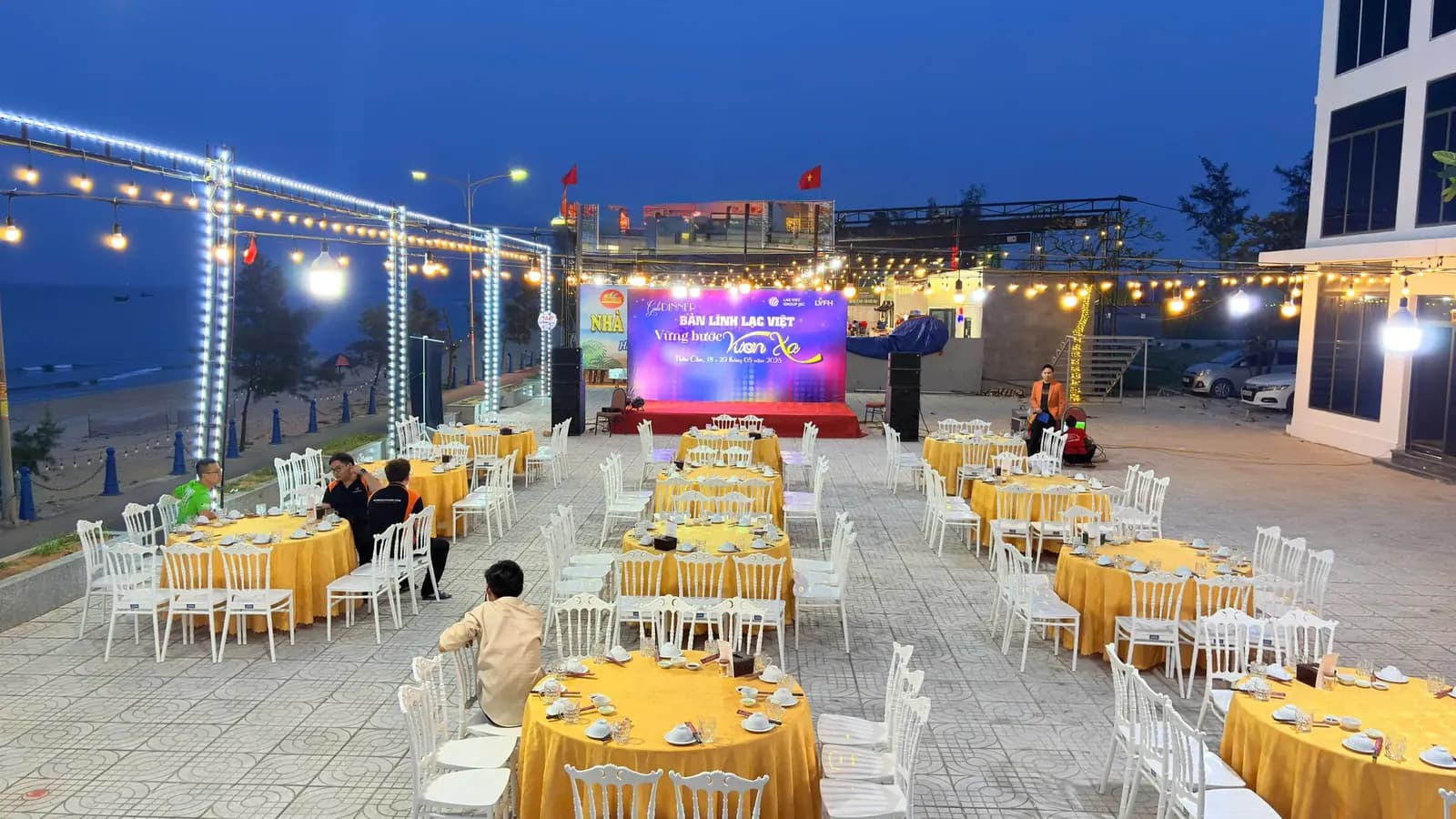 Khu vực gala dinner ngoài trời đang được setup buổi chiều: bàn ghế bày sẵn, sân khấu LED và hệ thống đèn dây hướng ra biển Thiên Cầm